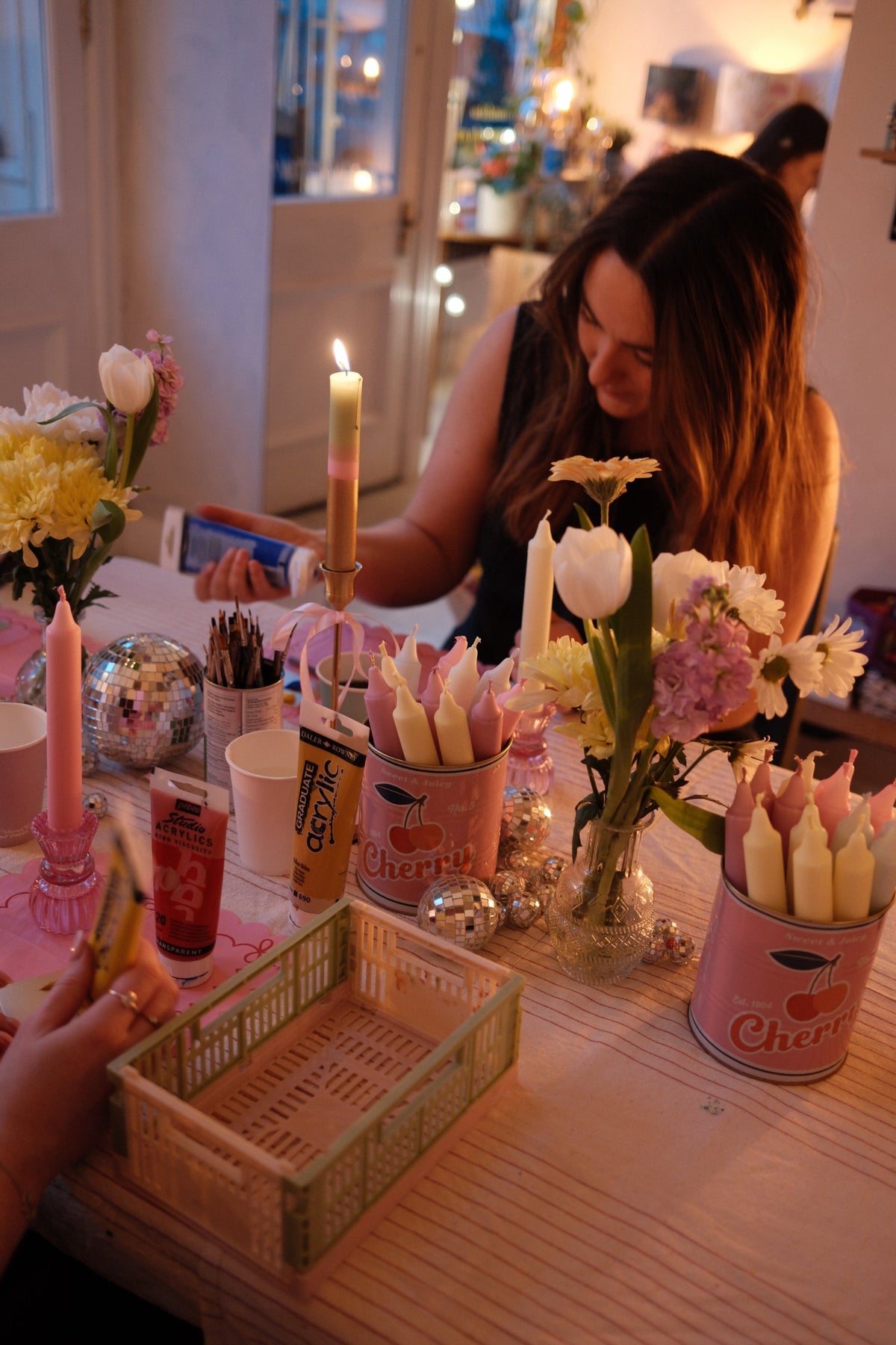 Person decorating a candle with flowers and a cans of candles on a table.  A lit pink and green candle is in the centre of the photo