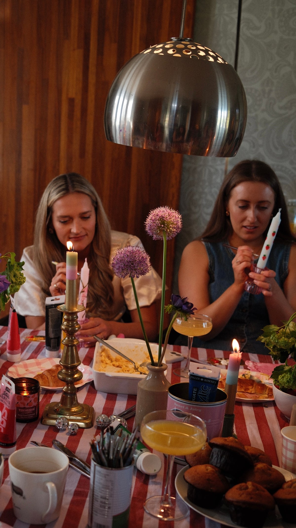 Two women sitting at a table with food and drinks, each decorating a candle. A Cocktail is at the forefront of the picture