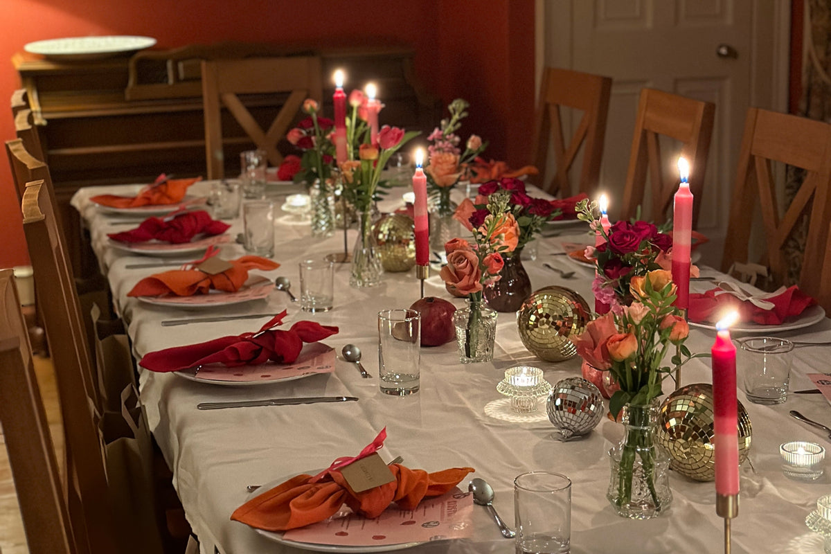 A dining room setting with dark pink and pink candles which are lit, small bud roses in glass jars, mirror balls, and autumnal coloured cloth napkins all on a white tablecloth